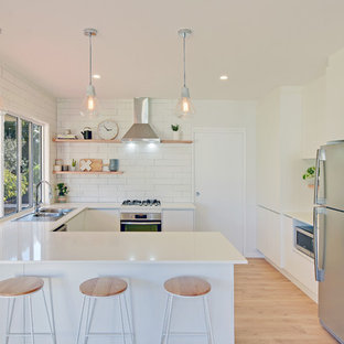 Design ideas for a beach style u-shaped separate kitchen in Sunshine Coast with a drop-in sink, flat-panel cabinets, white cabinets, white splashback, subway tile splashback, stainless steel appliances, light hardwood floors, a peninsula and beige floor.