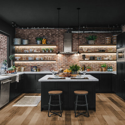 Example of a large urban u-shaped laminate floor and brown floor kitchen design in Los Angeles with a farmhouse sink, shaker cabinets, black cabinets, red backsplash, brick backsplash, stainless steel appliances, an island and white countertops