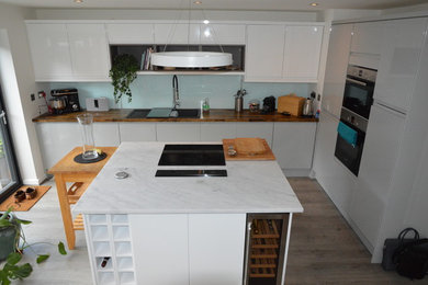 This is an example of a large modern l-shaped open plan kitchen in Manchester with a double-bowl sink, flat-panel cabinets, white cabinets, composite countertops, blue splashback, glass sheet splashback, black appliances, medium hardwood flooring, an island and grey floors.