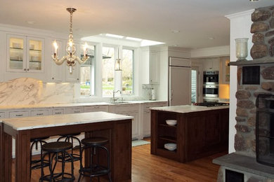 Example of a large classic l-shaped light wood floor eat-in kitchen design in New York with a single-bowl sink, recessed-panel cabinets, white cabinets, marble countertops, white backsplash, stone slab backsplash, stainless steel appliances and two islands