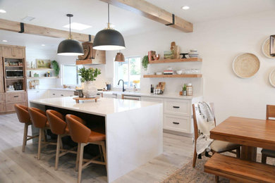 Example of a large minimalist single-wall laminate floor, gray floor and exposed beam eat-in kitchen design in Other with a farmhouse sink, shaker cabinets, white cabinets, quartz countertops, white backsplash, quartz backsplash, white appliances, an island and white countertops