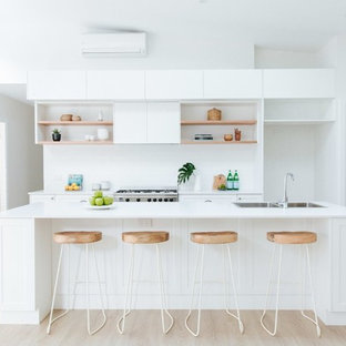 Photo of a beach style galley kitchen in Wollongong with a double-bowl sink, shaker cabinets, white cabinets, white splashback, stainless steel appliances, light hardwood floors, with island, beige floor and white benchtop.