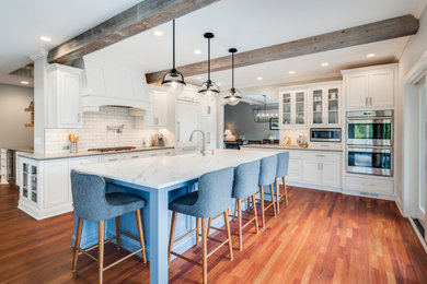 Large transitional l-shaped medium tone wood floor and brown floor kitchen photo in Minneapolis with a farmhouse sink, recessed-panel cabinets, white cabinets, white backsplash, subway tile backsplash, stainless steel appliances, an island and white countertops