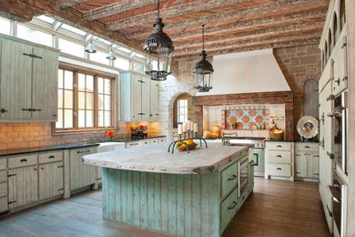 Large mountain style u-shaped medium tone wood floor and brown floor kitchen photo in Dallas with a farmhouse sink, flat-panel cabinets, distressed cabinets, solid surface countertops, beige backsplash, stone tile backsplash, stainless steel appliances and an island