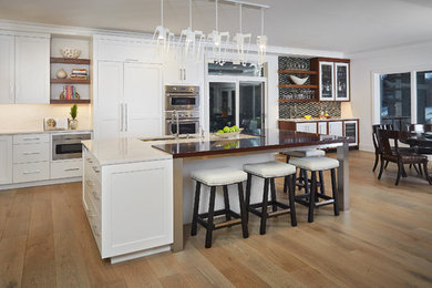 Open concept kitchen - large transitional l-shaped medium tone wood floor open concept kitchen idea in Grand Rapids with an undermount sink, recessed-panel cabinets, white cabinets, wood countertops, white backsplash, stainless steel appliances and an island