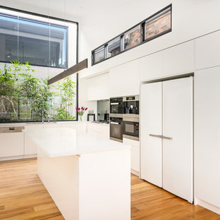 Mid-sized contemporary l-shaped kitchen in Sydney with a drop-in sink, flat-panel cabinets, white cabinets, stainless steel appliances, medium hardwood floors, multiple islands, brown floor, white benchtop, grey splashback and glass sheet splashback.