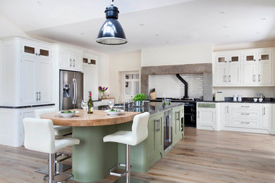 This is an example of a large farmhouse l-shaped kitchen/diner in Belfast with a submerged sink, shaker cabinets, green cabinets, granite worktops, white splashback, ceramic splashback, stainless steel appliances, light hardwood flooring, an island and brown floors.