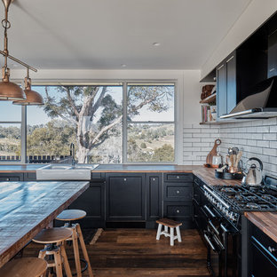 Design ideas for a country l-shaped kitchen in Sunshine Coast with a farmhouse sink, recessed-panel cabinets, black cabinets, wood benchtops, white splashback, subway tile splashback, black appliances, dark hardwood floors, with island, brown floor and brown benchtop.