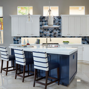 Photo of a large beach style l-shaped open plan kitchen in Miami with an undermount sink, shaker cabinets, white cabinets, blue splashback, glass tile splashback, stainless steel appliances, marble floors, with island and beige floor.