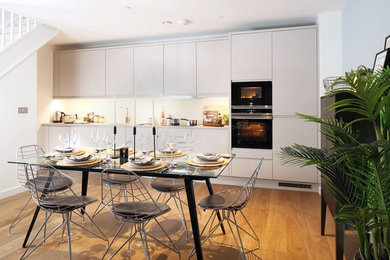 Inspiration for a medium sized modern single-wall kitchen/diner in London with a single-bowl sink, flat-panel cabinets, white cabinets, laminate countertops, multi-coloured splashback, glass sheet splashback, white appliances, light hardwood flooring, multiple islands, brown floors and white worktops.