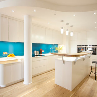 Inspiration for a mid-sized scandinavian l-shaped kitchen in London with flat-panel cabinets, white cabinets, glass benchtops, blue splashback, glass sheet splashback, light hardwood floors and with island.