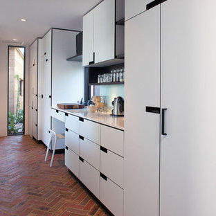 Modelo de cocina de galera, minimalista, con despensa, fregadero encastrado, armarios con paneles lisos, puertas de armario blancas, encimera de granito, salpicadero blanco, salpicadero de vidrio templado, electrodomésticos negros, suelo de ladrillo y una isla