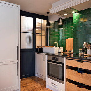 Small contemporary l-shaped kitchen/diner in London with a single-bowl sink, flat-panel cabinets, beige cabinets, granite worktops, green splashback, ceramic splashback, stainless steel appliances, light hardwood flooring, no island, brown floors and black worktops.