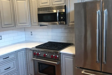 Example of a mid-sized transitional l-shaped medium tone wood floor and brown floor kitchen design in DC Metro with raised-panel cabinets, gray cabinets, quartz countertops, white backsplash, subway tile backsplash, stainless steel appliances and white countertops