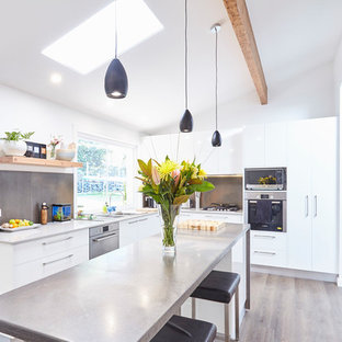 Inspiration for a contemporary l-shaped kitchen in Central Coast with a double-bowl sink, flat-panel cabinets, white cabinets, stainless steel benchtops, grey splashback, black appliances, light hardwood floors, with island, brown floor and grey benchtop.