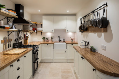 Kitchen - small transitional u-shaped travertine floor and beige floor kitchen idea in London with a farmhouse sink, shaker cabinets, white cabinets, wood countertops, white backsplash, subway tile backsplash, black appliances and brown countertops