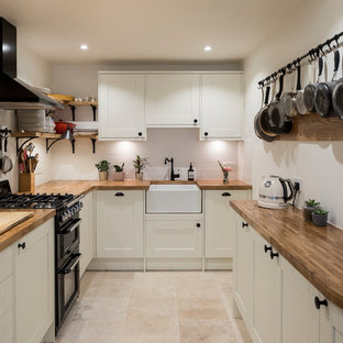 Small traditional u-shaped kitchen in London with a belfast sink, shaker cabinets, white cabinets, wood worktops, white splashback, metro tiled splashback, black appliances, travertine flooring, beige floors and brown worktops.