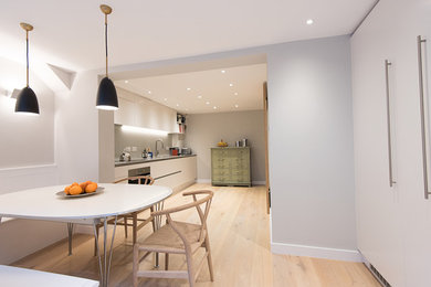 Photo of a contemporary kitchen/diner in London with a double-bowl sink, flat-panel cabinets, white cabinets, engineered stone countertops, grey splashback, glass sheet splashback, stainless steel appliances and light hardwood flooring.