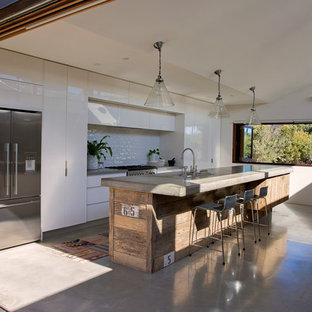 Photo of a large beach style galley open plan kitchen in Sydney with a drop-in sink, flat-panel cabinets, white cabinets, concrete benchtops, white splashback, subway tile splashback, stainless steel appliances, concrete floors, with island, grey floor and grey benchtop.