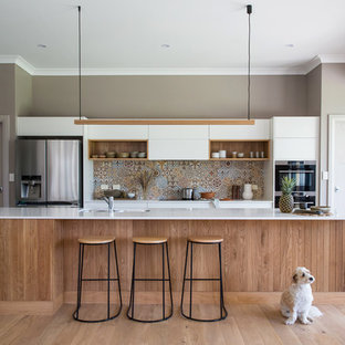 This is an example of a large transitional galley kitchen in Other with a double-bowl sink, flat-panel cabinets, white cabinets, solid surface benchtops, multi-coloured splashback, ceramic splashback, stainless steel appliances, light hardwood floors, with island, brown floor and white benchtop.