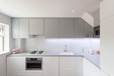 This is an example of a small contemporary l-shaped open plan kitchen in London with a built-in sink, flat-panel cabinets, white cabinets, composite countertops, white splashback, mosaic tiled splashback, stainless steel appliances, dark hardwood flooring and an island.