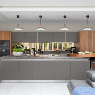 Large contemporary galley open plan kitchen in Hampshire with grey cabinets, quartz worktops, metallic splashback, mirror splashback, black appliances, an island, grey worktops, flat-panel cabinets and grey floors.