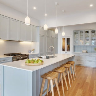 Mid-sized beach style kitchen in Sydney with a single-bowl sink, shaker cabinets, grey cabinets, solid surface benchtops, white splashback, stainless steel appliances, with island, white benchtop, light hardwood floors and beige floor.