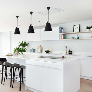 Contemporary galley kitchen in London with a submerged sink, flat-panel cabinets, white cabinets, black appliances, light hardwood flooring, an island, beige floors and white worktops.