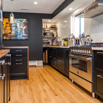 Enclosed kitchen - mid-sized contemporary galley light wood floor and beige floor enclosed kitchen idea in New York with an undermount sink, recessed-panel cabinets, black cabinets, wood countertops, white backsplash, stainless steel appliances, an island, brown countertops and subway tile backsplash