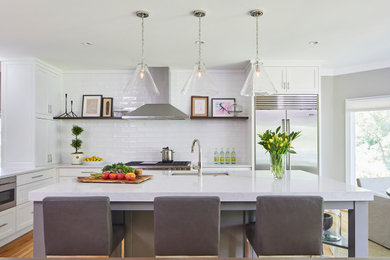 Diseño de cocina comedor actual grande con fregadero bajoencimera, armarios estilo shaker, puertas de armario blancas, encimera de mármol, salpicadero blanco, salpicadero de azulejos de cerámica, electrodomésticos de acero inoxidable, suelo de madera en tonos medios, una isla y encimeras blancas