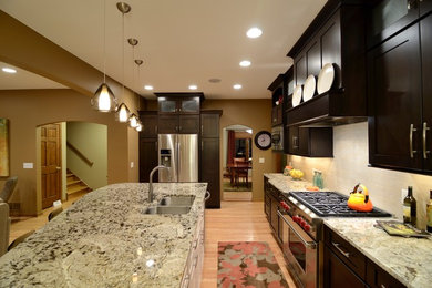 Mid-sized trendy galley light wood floor open concept kitchen photo in Minneapolis with an undermount sink, flat-panel cabinets, dark wood cabinets, granite countertops, white backsplash, stone tile backsplash, stainless steel appliances and an island