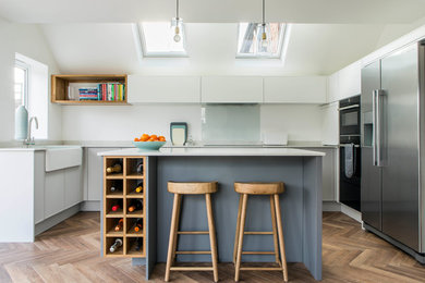 Design ideas for a contemporary kitchen in Oxfordshire with an island, a belfast sink, flat-panel cabinets, white cabinets, glass sheet splashback, black appliances, light hardwood flooring and grey worktops.