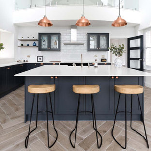 Photo of a large classic l-shaped kitchen in Essex with a submerged sink, shaker cabinets, black cabinets, composite countertops, white splashback, ceramic splashback, integrated appliances, light hardwood flooring, an island, beige floors and white worktops.