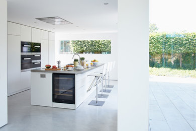 Modern inredning av ett stort linjärt kök och matrum, med en köksö, släta luckor, vita skåp, bänkskiva i rostfritt stål, svarta vitvaror, en integrerad diskho och betonggolv