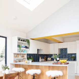 Large contemporary u-shaped kitchen/diner in Hertfordshire with flat-panel cabinets, white cabinets, composite countertops, blue splashback, integrated appliances, light hardwood flooring, a breakfast bar, beige floors and white worktops.