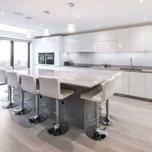 Inspiration for an expansive modern single-wall open plan kitchen in London with flat-panel cabinets, grey cabinets, quartz worktops, white splashback, glass sheet splashback, black appliances, an island, grey worktops, a submerged sink, light hardwood flooring and beige floors.