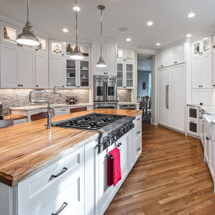 Large transitional u-shaped medium tone wood floor and brown floor enclosed kitchen photo in San Diego with a farmhouse sink, shaker cabinets, white cabinets, wood countertops, gray backsplash, stone tile backsplash, stainless steel appliances, an island and white countertops