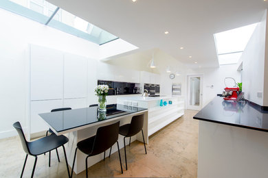 This is an example of a large galley open plan kitchen in Other with a double-bowl sink, flat-panel cabinets, white cabinets, composite countertops, black splashback, glass sheet splashback, stainless steel appliances, limestone flooring and an island.
