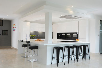Пример оригинального дизайна: кухня-гостиная среднего размера в современном стиле с двойной мойкой, плоскими фасадами, белыми фасадами, столешницей из акрилового камня, серым фартуком, черной техникой и островом