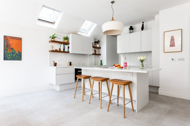 This is an example of a medium sized contemporary kitchen/diner in London with a built-in sink, flat-panel cabinets, white cabinets, marble worktops, white splashback, marble splashback, black appliances, ceramic flooring, an island, white floors and white worktops.
