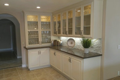 Example of a mid-sized trendy l-shaped ceramic tile enclosed kitchen design in Los Angeles with a farmhouse sink, stainless steel appliances, white backsplash, stone tile backsplash, raised-panel cabinets, white cabinets, solid surface countertops and an island