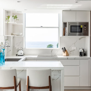 This is an example of a beach style u-shaped kitchen in Sydney with a drop-in sink, flat-panel cabinets, white cabinets, white splashback, stainless steel appliances, beige floor and white benchtop.