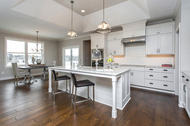 Inspiration for a transitional l-shaped medium tone wood floor and brown floor open concept kitchen remodel in Other with a farmhouse sink, shaker cabinets, white cabinets, granite countertops, white backsplash, subway tile backsplash, stainless steel appliances and an island