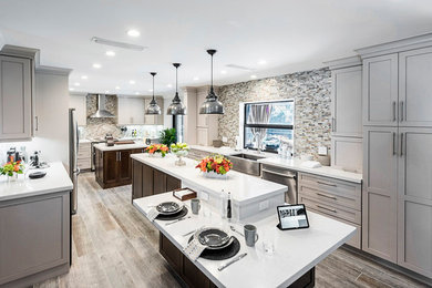 Large traditional galley enclosed kitchen in Miami with a belfast sink, shaker cabinets, beige cabinets, engineered stone countertops, beige splashback, stone tiled splashback, stainless steel appliances, light hardwood flooring, multiple islands and grey floors.