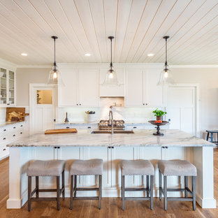 Example of a classic l-shaped medium tone wood floor and brown floor kitchen design in Other with a farmhouse sink, shaker cabinets, white cabinets, white backsplash, subway tile backsplash, stainless steel appliances, an island and gray countertops