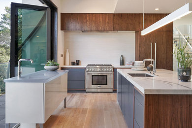 Example of a trendy light wood floor and beige floor kitchen design in San Francisco with an undermount sink, flat-panel cabinets, gray cabinets, white backsplash, stainless steel appliances, two islands and white countertops
