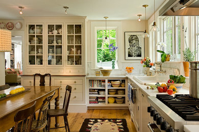 Example of a classic eat-in kitchen design in Burlington with a drop-in sink, glass-front cabinets, beige cabinets, white backsplash, stone slab backsplash and stainless steel appliances