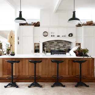 Inspiration for a large country galley kitchen in Sydney with a farmhouse sink, shaker cabinets, white cabinets, quartz benchtops, grey splashback, stone tile splashback, black appliances, travertine floors, with island, grey floor and white benchtop.