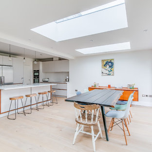 Medium sized contemporary l-shaped kitchen/diner in London with a submerged sink, flat-panel cabinets, white cabinets, composite countertops, white splashback, stainless steel appliances, an island, beige floors, light hardwood flooring and white worktops.