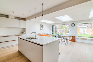 Inspiration for a medium sized contemporary l-shaped kitchen/diner in Surrey with a submerged sink, flat-panel cabinets, white cabinets, quartz worktops, white splashback, stainless steel appliances, light hardwood flooring and an island.